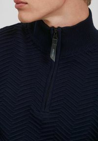 Pullover blu navy con colletto alto a zip, caratterizzato da un motivo a zigzag testurizzato. La zip è dotata di un tirante nero etichettato "JEANS CODE."