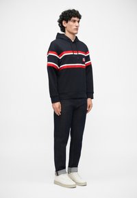 Sudadera azul marino con franjas horizontales rojas y blancas, bolsillo frontal, capucha con cordón. Combinada con jeans oscuros y zapatillas blancas.