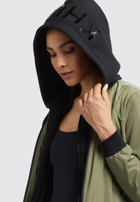 Jachetă bomber de culoare verde măslin cu glugă neagră, având inscriptionări lucioase, manșete strâmte și închidere cu fermoar. Textură netedă și design ajustat.