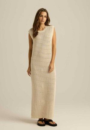 Dzianinowa długa sukienka w kolorze kremowym o luźnym kroju, z poziomymi i pionowymi ażurowymi wzorami oraz okrągłym dekoltem. Zestawiona z czarnymi sandałami.