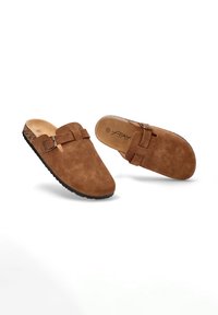 Zuecos de ante marrón sin hebilla, con una correa y cierre prominentes. Suela texturizada para un buen agarre, diseño de punta curva y plantilla de madera natural.