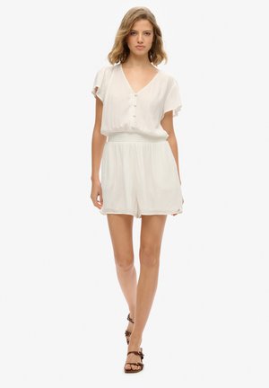 Junge Frau geht vorwärts, trägt einen weißen kurzärmeligen Overall und braune Sandalen mit Absatz vor einem einfarbigen Hintergrund.