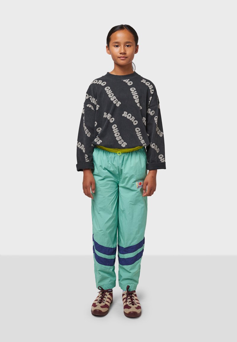 Schwarzes Langarmshirt mit wiederholtem Text "BOBO CHOSES". Hellgrüne Hose mit Kordelzug und blauen Streifen. Braune und cremfarbene Schuhe.