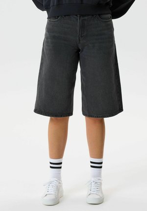 Person trägt schwarze kniehohe Jeansshorts, weiße Socken mit schwarzen Streifen und weiße Turnschuhe, steht vor weißem Hintergrund.