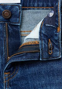 Džins hlače v temno modri barvi z vidnim šivanjem, kovinsko gumbom, zadrgo in odprtino za žep, ki razkriva svetlejšo notranjo tkanino.