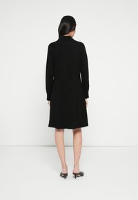 Robe noire à manches longues avec un motif texturé, coupe jusqu'aux genoux, et un col simple. Portée avec des talons ouverts noirs.