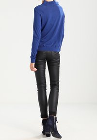 Donna che indossa un maglione blu, pantaloni di pelle nera e stivaletti con tacco blu scuro in piedi su uno sfondo bianco uniforme.