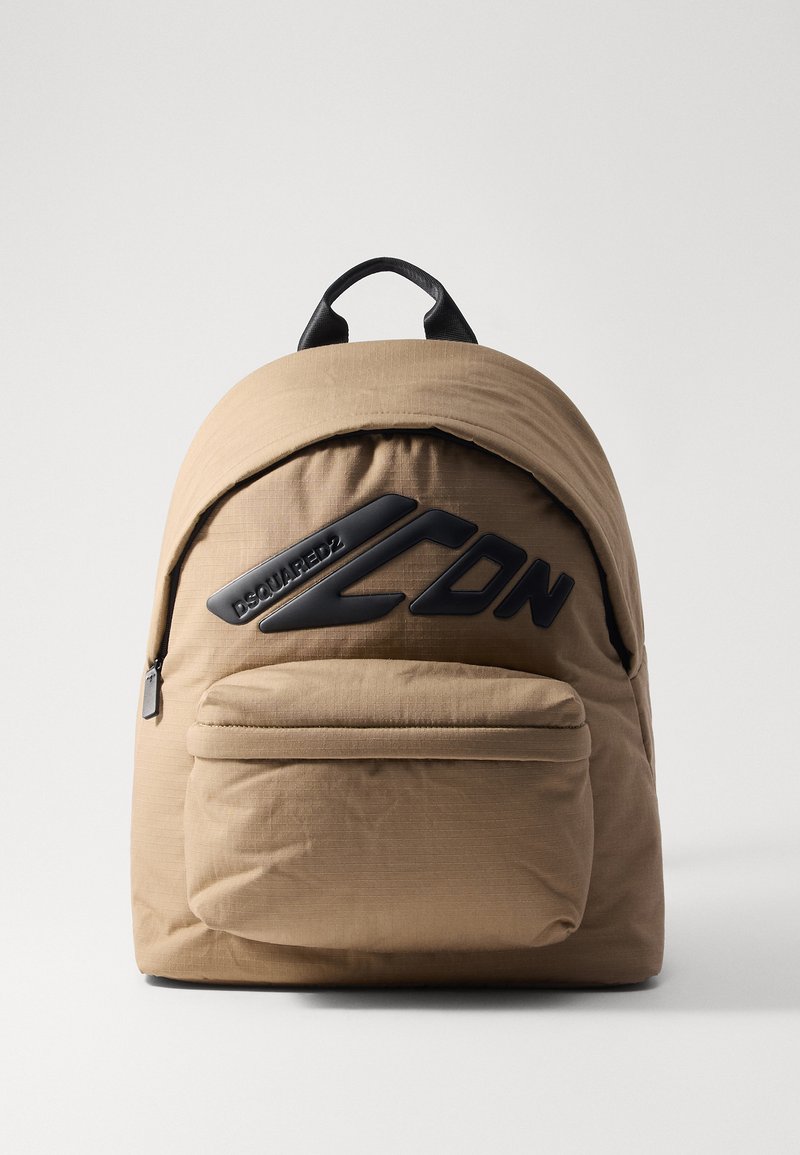 Tan nylon rygsæk med rundetop, stor frontlomme og sorte logo-detaljer. Har dobbelte top-håndtag og en tekstureret overflade.
