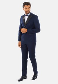Giacca da smoking doppiopetto blu navy con rever in raso, abbinata a pantaloni coordinati e scarpe eleganti nere lucide. Camicia bianca e papillon.