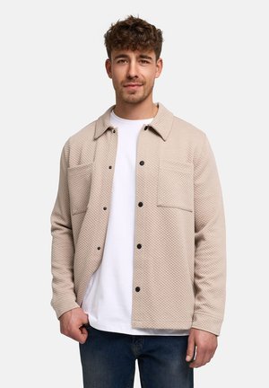 Ung mand med kort krøllet hår iført en beige quiltet skjortejakke med knapper over en hvid T-shirt og blå jeans, stående mod en ensfarvet baggrund.