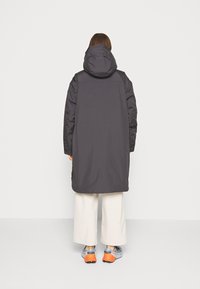 Parka larga de un gris oscuro con capucha, con una textura suave y aberturas laterales, combinada con pantalones de pierna ancha color crema y deportivas gris-naranja.