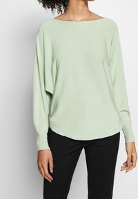 Suéter de gran tamaño en verde claro, de tejido de punto suave, con escote barco ancho, mangas largas y dobladillo curvado. Combinado con pantalones negros.