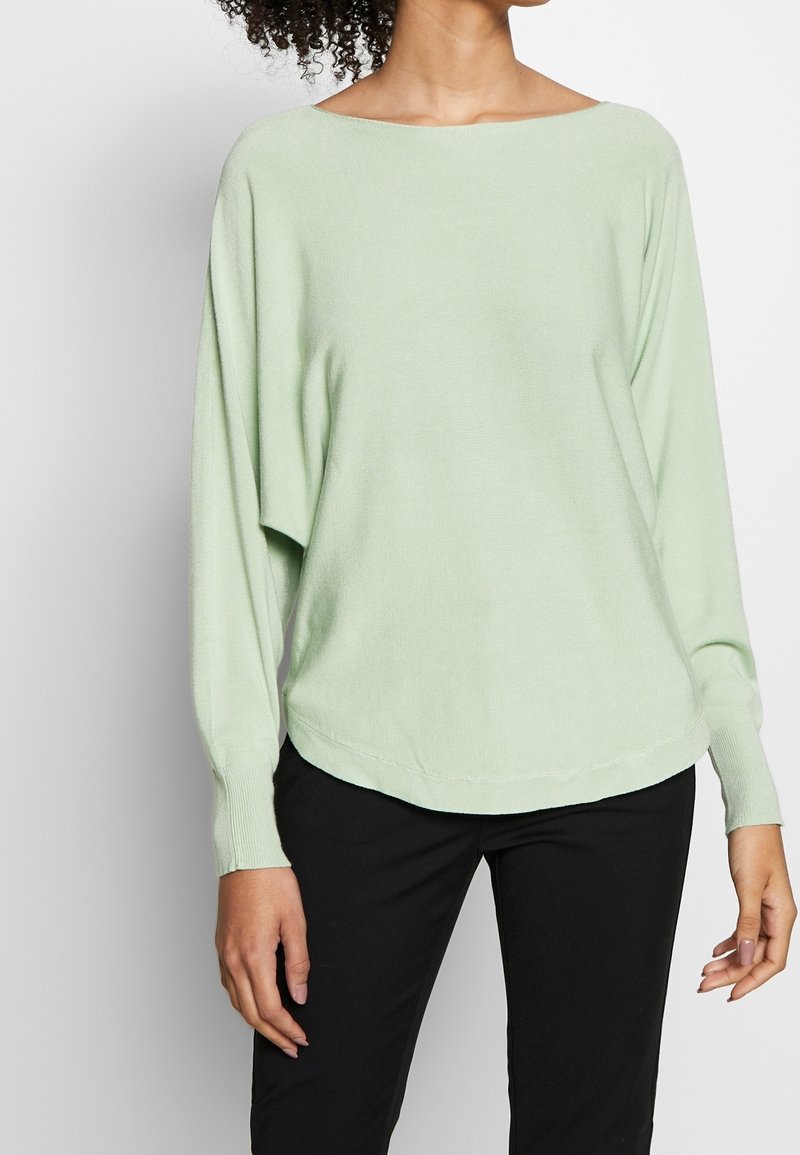 Suéter de gran tamaño en verde claro, de tejido de punto suave, con escote barco ancho, mangas largas y dobladillo curvado. Combinado con pantalones negros.