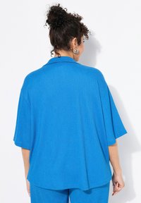 Camicia oversize blu con un tessuto texturizzato, maniche corte e colletto. Il retro presenta un orlo dritto e una vestibilità comoda.
