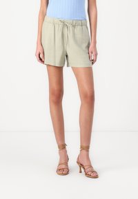 Shorts vert clair avec une taille élastique et un cordon de serrage, accompagnés de talons beiges à lanières. Texture lisse, design décontracté.