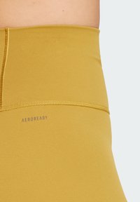 Leggings taille haute jaunes AEROREADY, fabriqués en tissu lisse avec un léger éclat, présentant des coutures ton sur ton et un logo imprimé.