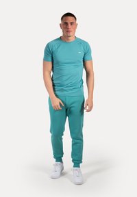 Top deportivo en turquesa y pantalones de chándal a juego. Hecho de una tela suave, con cuello redondo y puños elásticos. Zapatillas blancas completan el conjunto.