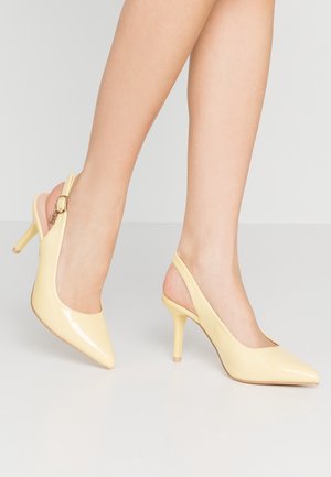 Benen met lichtgele lakleren slingback hoge hakken met puntige neuzen en smalle hakken op een lichtgrijze vloer.