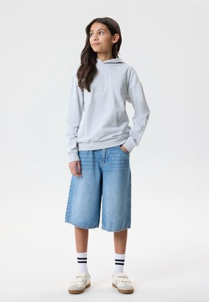 Pige iført lys grå hættetrøje, knælange denimshorts, hvide sokker med sorte striber og hvide sneakers, stående med den ene hånd i lommen.