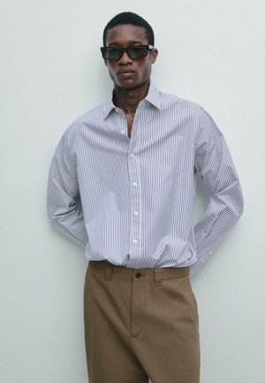 Mann mit schwarzen Sonnenbrillen, einem weißen Hemd mit dünnen vertikalen Streifen und brauner Hose, der vor einer schlichten hellen Wand steht.