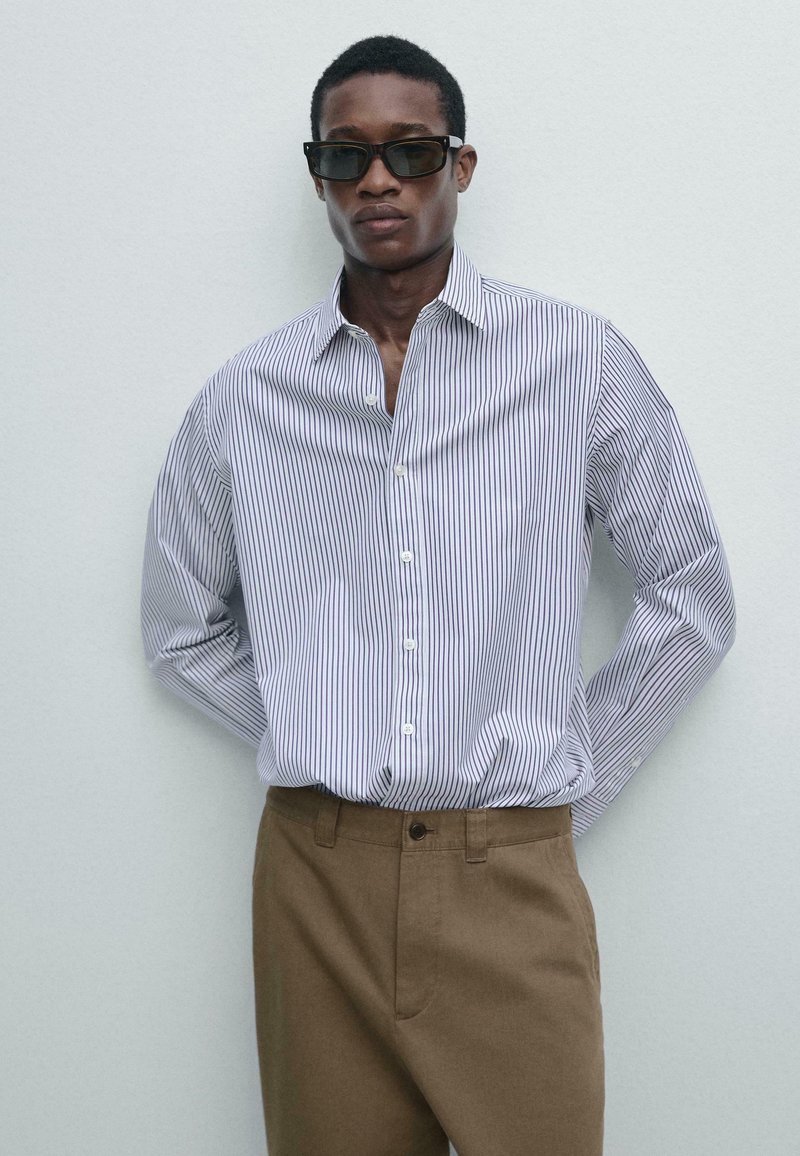 Mand iført sorte solbriller, en hvid skjorte med tynde lodrette striber og brune bukser, stående foran en ensfarvet lys væg.