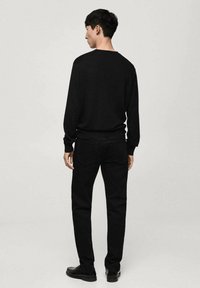 Maglione nero a maniche lunghe con polsini a coste, abbinato a jeans neri slim-fit. Il modello sta di spalle, mostrando un design minimale.