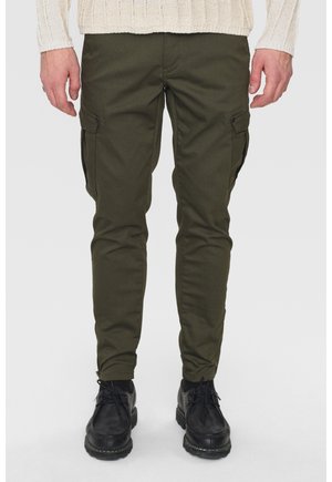 Mężczyzna ubrany w oliwkowozielone obcisłe spodnie typu cargo, czarne sznurowane buty oraz kremowy sweter w warkocze, stojący na tle jednolitego tła.