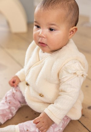 Fleece-Creme-Weste mit zwei braunen Knöpfen, über einem hellen, strukturierten Langarmshirt getragen. Weiche, rosa gemusterte Hose mit Vögeln.