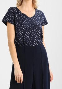 Top azul marino con escote en V y mangas cortas, con un estampado de corazones blancos dispersos. Tejido suave y corte relajado. Combinado con pantalones de tiro alto.