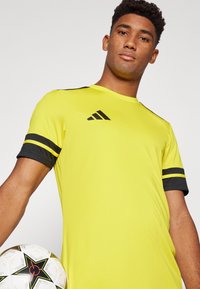 Yellow sports jersey with black accents and stripes, featuring the Adidas logo on the chest. Player holds a soccer ball with a star pattern.