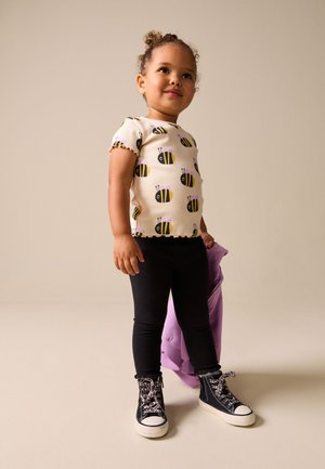 Fetiță mică purtând un tricou cu imprimeu cu albine, colanți negri și adidași înalți, ținând o geacă violet pliată.