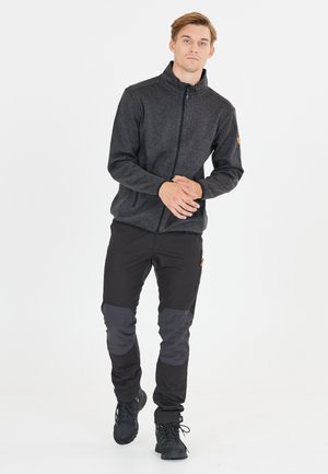 Veste noire zippée pour homme en tissu texturé, associée à un pantalon noir avec des panneaux gris aux genoux et des chaussures de randonnée noires.