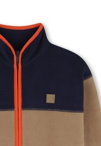 Giacca in pile con parte superiore blu navy, parte inferiore beige e cerniera arancione. Presenta una piccola etichetta con logo e cuciture a contrasto. Design con colletto alto.