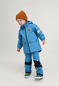 Bambino con giacca e pantaloni impermeabili blu, cappello in maglia arancione e stivali da pioggia verdi in piedi davanti a uno sfondo semplice.