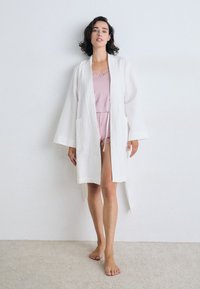 Hvit teksturert morgenkåpe med brede ermer og sidelommer, lagd over et rosa camiskjorte- og shorts-sett med blondedetaljer. Mykt, lett stoff.