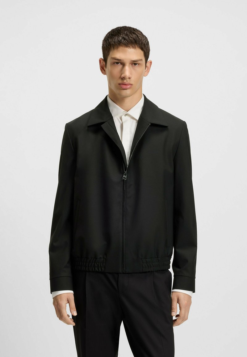 Black zip-up jacket with a collared design, elastic waistband, and smooth, lightweight fabric. Features side pockets and tailored fit.