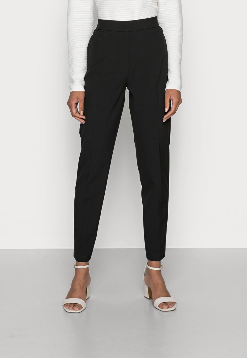 Žena oblečená v černých vysokých tapered kalhotách, bílém ribaném topu s dlouhým rukávem a bílých otevřených kotníkových sandálech na bloku stojí na světlé podlaze.