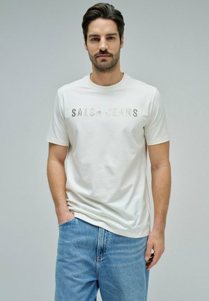 Homme aux cheveux foncés et à la barbe portant un t-shirt blanc "SALSA JEANS" et un jean bleu, debout devant un fond gris clair uni.