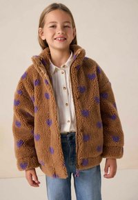 Veste en polaire marron avec des motifs de cœurs violets, fermeture éclair et poches latérales. Portée par-dessus une chemise blanche à boutons et un jean bleu.