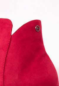 Détail d'une bottine en daim rouge montrant une texture lisse et un petit emblème métallique rond près du bord supérieur.