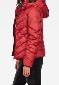 Doudoune rouge avec capuche, présentant un design en chevrons matelassé, une fermeture éclair et un écusson circulaire sur la manche gauche. Texture douce.