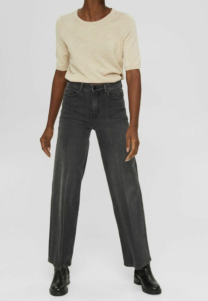 Haut en maille à manches courtes de couleur beige associé à un jean taille haute, coupe large, gris foncé, et des bottines noires. Tissu lisse avec texture classique du denim.