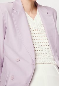 Jasnofioletowa marynarka z dwiema dużymi guzikami, klapami i gładkim materiałem, narzucona na białą swetrową bluzkę o fakturze.