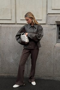 Bruine nepleer oversized jas met een grote kraag, gecombineerd met een paar bruine broeken en witte flats, terwijl ze een kleine witte handtas vasthoudt.