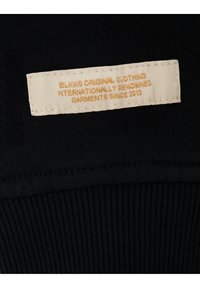 Sort ribbet stof med et beige tekstilmærke syet på, der har præget guldtekst: "BLKWS ORIGINAL CLOTHING INTERNATIONALLY RENOWNED GARMENTS SINCE 2013."