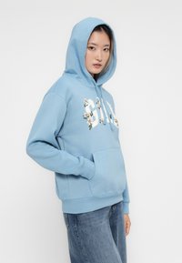 Light blue hoodie with a front pocket, featuring floral designs and bold lettering. Soft texture, relaxed fit, and drawstring hood.