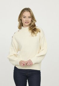 Pull en maille couleur crème avec un col montant, un motif texturé et des manches bouffantes. Présente des boutons décoratifs sur l'épaule.