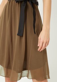 Vestido de gasa marrón con un cinturón de lazo negro. La tela es ligera y semitranslúcida, con una silueta fluida y un dobladillo en capas.