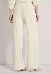 Vrouw in hoge taille crêpe brede broek en beige puntige hakken, staande tegen een effen achtergrond.