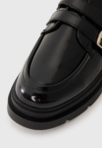 Black patent leather ankle boot with a rounded toe, two adjustable straps, and a chunky black sole. Features subtle stitching details.